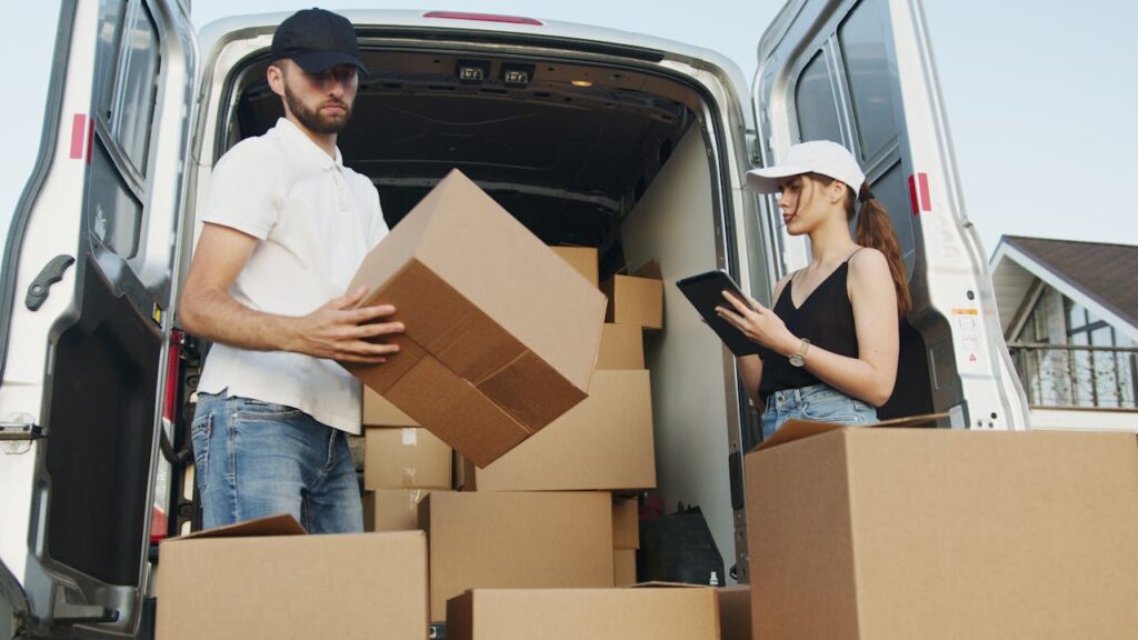 pexels photo 5025667 Two delivery workers handling cardboard boxes by a van, checking orders on tablet.